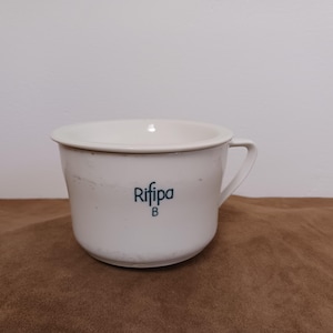 Könnte beinhalten: Weiße Keramik-Nachttopf mit Henkel. Der Topf hat einen breiten Rand und einen abgerundeten Körper. Der Text "Rifipa" ist in Dunkelblau auf die Vorderseite gedruckt, darunter ein "B". Der Topf weist Gebrauchsspuren auf.