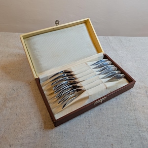 Könnte beinhalten: Ein Set silberfarbener Gabeln in einer braunen und beigefarbenen Box. Die Gabeln haben ein einzigartiges, geschwungenes Design. Die rechteckige Box hat einen Klappdeckel und einen kleinen Verschluss. Der Innenraum ist mit cremefarbenem Stoff ausgekleidet.