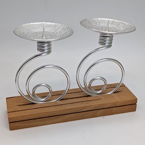 Könnte beinhalten: Zwei silberfarbene Kerzenhalter mit Spiraldesign, jeweils mit einer strukturierten Schale und einem zentralen Dorn. Sie sind auf einem rechteckigen Holzsockel montiert und bilden eine dekorative und elegante Präsentation.