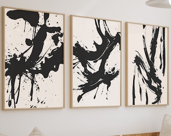 May include: A set of three framed wall art prints featuring bold black ink splatters and brushstrokes on a light cream background. Each rectangular print is encased in a thin, light-colored wooden frame, displayed side-by-side on a wall.