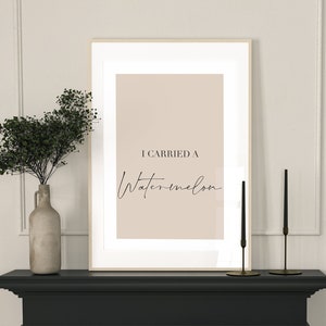 Peut inclure: Impression encadrée avec le texte « I Carried a Watermelon » dans un style minimaliste. L'impression est exposée sur une cheminée noire, à côté de bougeoirs noirs et d'un vase avec de la verdure. Une table à manger est dressée au premier plan avec des assiettes noires et de la verrerie.