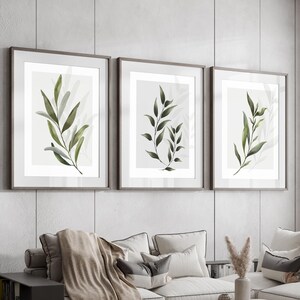 May include: A living room featuring a sectional sofa, three framed botanical prints, a coffee table, two ottomans, and a side table with a bookshelf. The sofa is upholstered in a light grey fabric and has a throw blanket draped over it. The prints depict simple line drawings of green leaves. The coffee table is made of wood and has a light grey finish. The ottomans are upholstered in a light grey fabric. The side table is made of dark wood and has a bookshelf on the bottom.