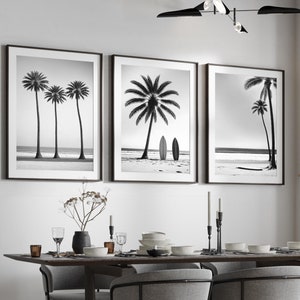 May include: Black and white photographs of palm trees on a beach, framed and hung on a white wall above a dining table with chairs. The table is set with plates, glasses, and cutlery.