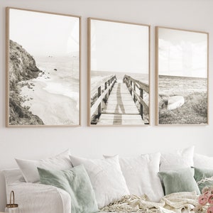 May include: Three black and white framed photographs of a beach scene with a wooden walkway leading to the ocean. The photographs are hanging on a white wall above a white couch with pillows and a throw blanket.