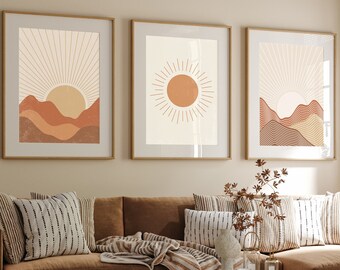 May include: Three framed wall art prints featuring abstract sun and mountain landscapes in shades of terracotta, beige, and cream. Each rectangular print is encased in a thin wooden frame, displaying geometric sunbursts and layered hills.