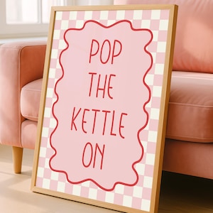 May include: A framed print featuring the text "POP THE KETTLE ON" in red on a pink, wavy-edged shape. The background has a pink and white checkered pattern. The frame is made of light wood.