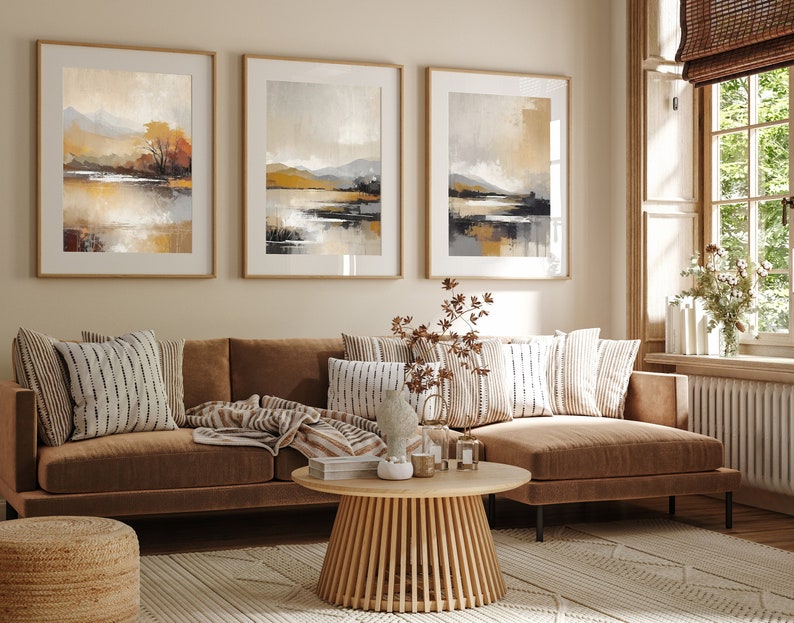 May include: A brown sectional sofa with a throw blanket and pillows in a living room with three framed abstract landscape paintings in muted tones of brown, beige, and gray. The sofa is in front of a window with a white windowsill and a brown window blind.