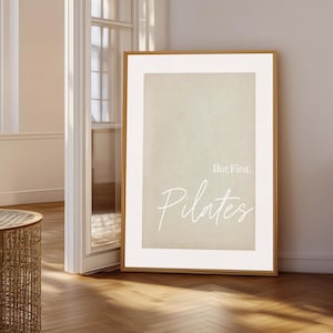 May include: A framed print with the text "But First, Pilates" in a white script font. The print has a neutral beige background and a wooden frame. The artwork is leaning against a wall in a room with a wooden floor.