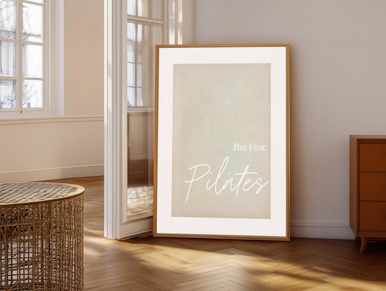 May include: A framed print with the text "But First, Pilates" in a white script font. The print has a neutral beige background and a wooden frame. The artwork is leaning against a wall in a room with a wooden floor.