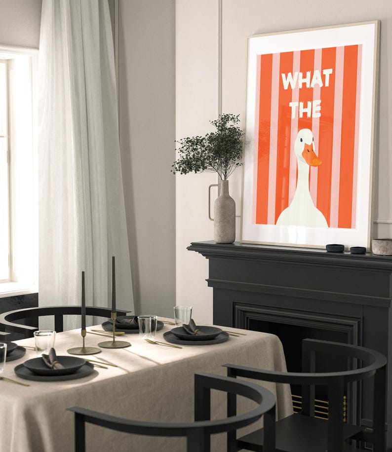 May include: A framed art print featuring a white goose with an orange beak against a red and white striped background. The text "WHAT THE" is displayed above the goose. The print is displayed above a black fireplace mantel.