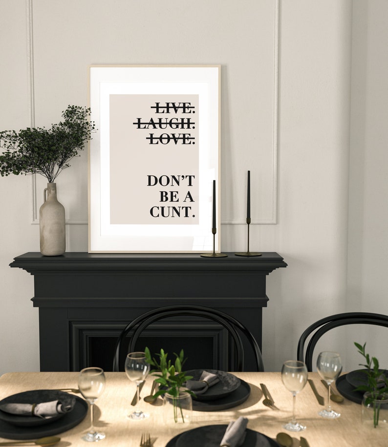 May include: A framed print with the text "LIVE. LAUGH. LOVE. DON'T BE A CUNT." hanging above a black fireplace mantel. The fireplace is decorated with two black candlesticks and a vase with greenery. The fireplace is in a dining room with a table set for dinner.