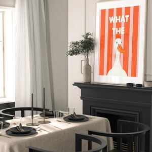 May include: A framed art print featuring a white goose with an orange beak against a red and white striped background. The text "WHAT THE" is displayed above the goose. The print is displayed above a black fireplace mantel.