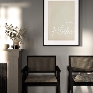 May include: A framed print with the text "But First, Pilates" in a white script font. The print is displayed above two black wooden chairs with woven seats and backs. A vase of dried flowers sits on a white mantel.