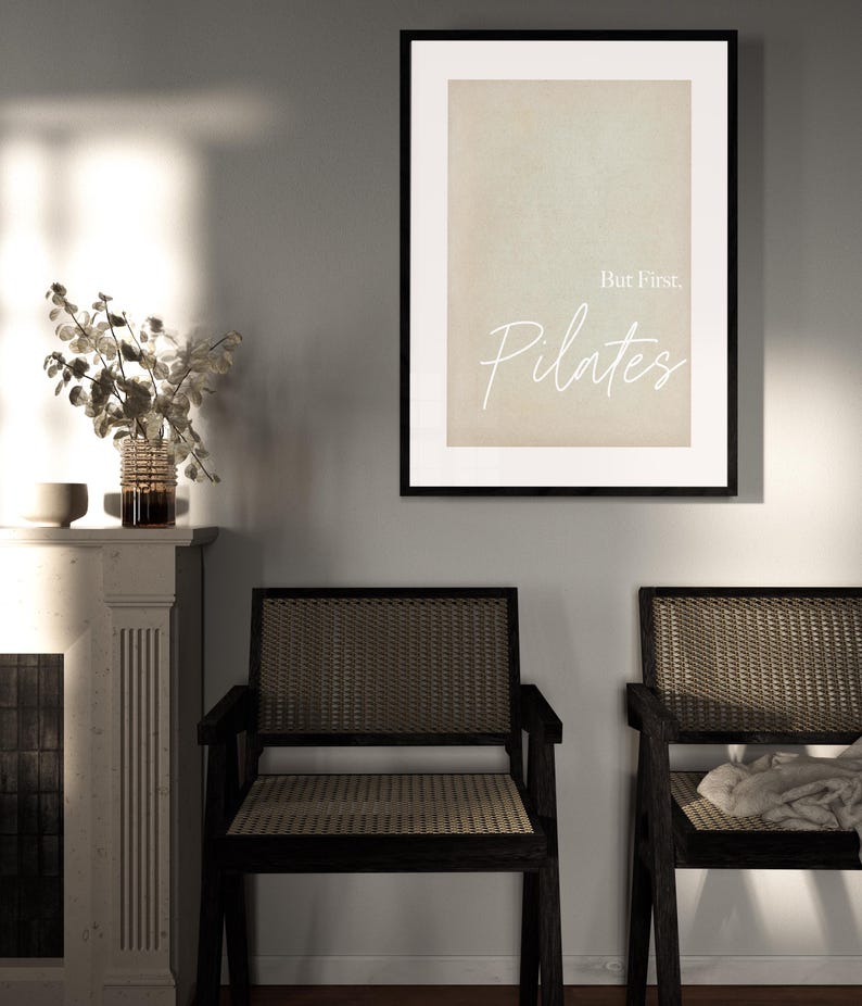 May include: A framed print with the text "But First, Pilates" in a white script font. The print is displayed above two black wooden chairs with woven seats and backs. A vase of dried flowers sits on a white mantel.