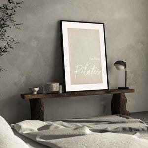 May include: A framed print with the text "But First, Pilates" in a minimalist style. The artwork is displayed on a rustic wooden shelf, accompanied by small decorative bowls and a modern black and white lamp. The background features a neutral-toned wall.