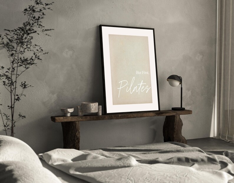 May include: A framed print with the text "But First, Pilates" in a minimalist style. The artwork is displayed on a rustic wooden shelf, accompanied by small decorative bowls and a modern black and white lamp. The background features a neutral-toned wall.