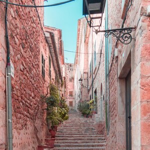 May include: A narrow, cobblestone street with pink stone buildings and a set of stairs leading up to the top. A black street lamp hangs from a wire above the stairs.