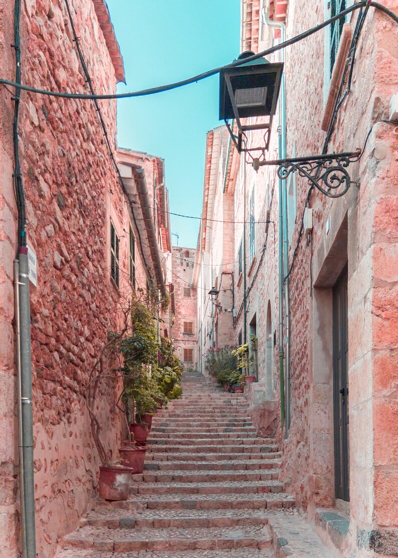May include: A narrow, cobblestone street with pink stone buildings and a set of stairs leading up to the top. A black street lamp hangs from a wire above the stairs.