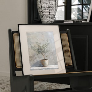 May include: A framed print of a watercolor painting of a small olive tree in a terracotta pot. The tree is green and the background is a soft blue and gray.