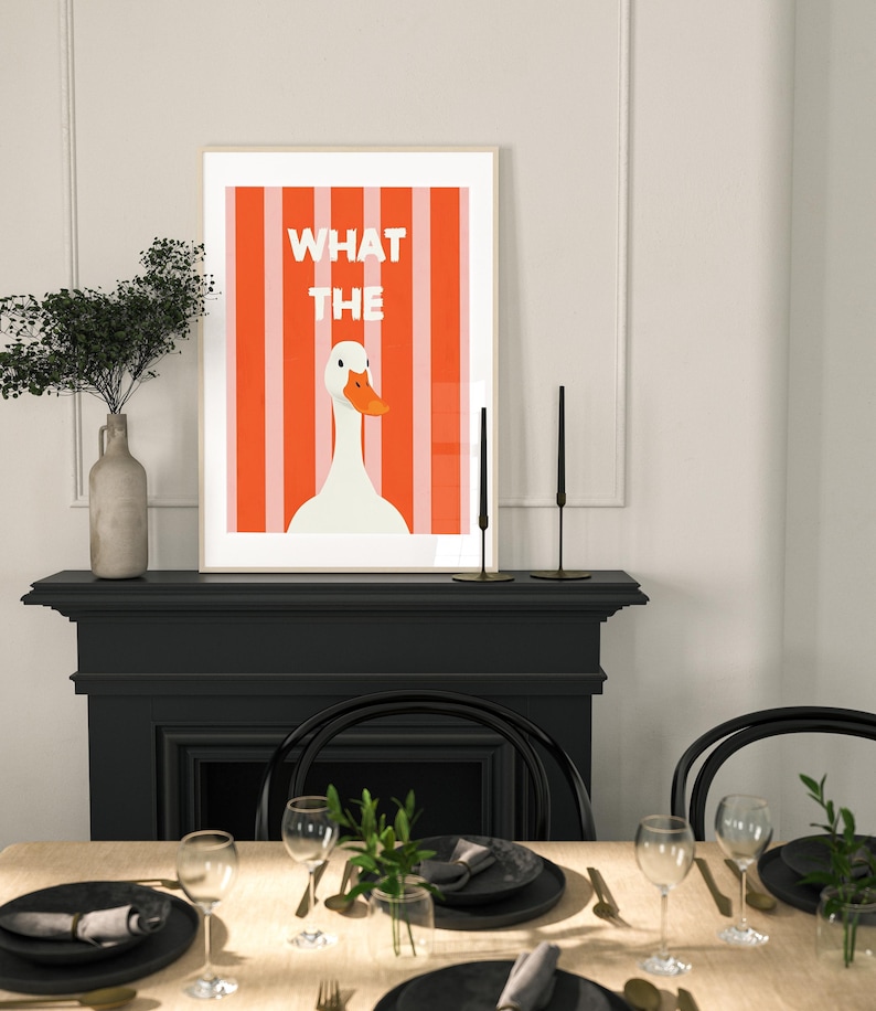 May include: A framed art print featuring a white goose with an orange beak against a background of orange and white stripes. The text "WHAT THE" is displayed above the goose. The print is displayed above a black fireplace mantel.