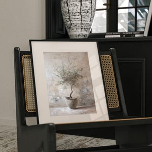 May include: A framed print of a watercolor painting of a small olive tree in a terracotta pot. The tree is in a light brown pot and is set against a light brown background with hints of green and blue.