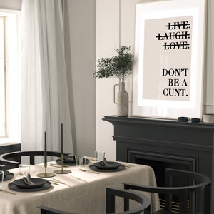 May include: A black framed poster with the text "LIVE. LAUGH. LOVE. DON'T BE A CUNT." hanging above a black fireplace mantel. The fireplace has a black hearth and a black firebox. A vase with dried flowers sits on the mantel. A dining table with a beige tablecloth and black chairs is in the foreground.