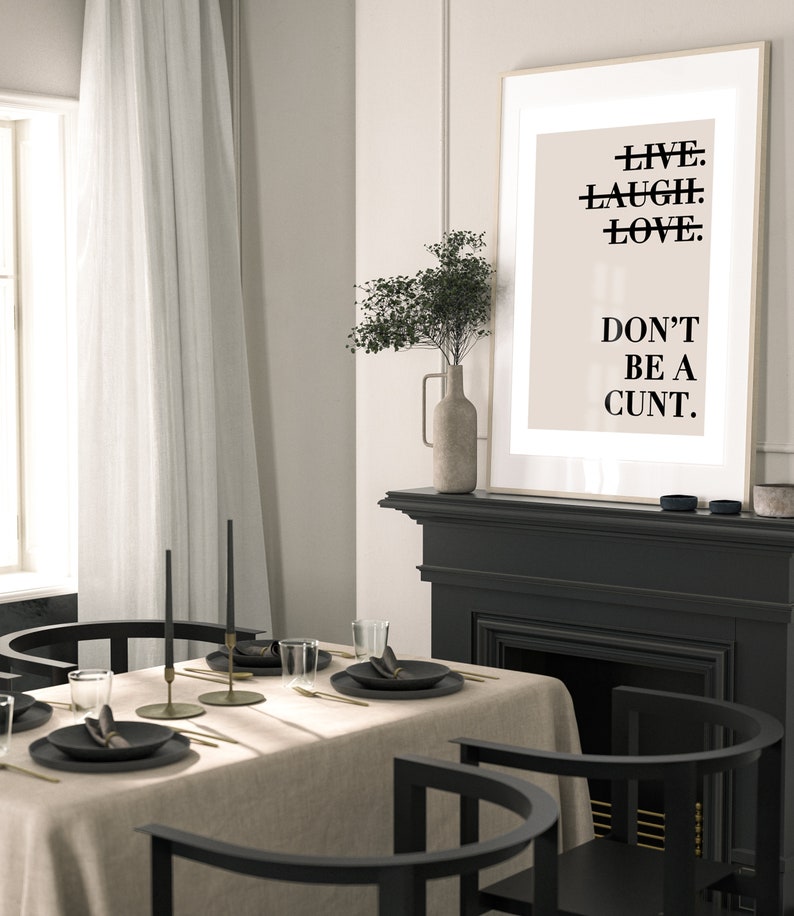 May include: A black framed poster with the text "LIVE. LAUGH. LOVE. DON'T BE A CUNT." hanging above a black fireplace mantel. The fireplace has a black hearth and a black firebox. A vase with dried flowers sits on the mantel. A dining table with a beige tablecloth and black chairs is in the foreground.
