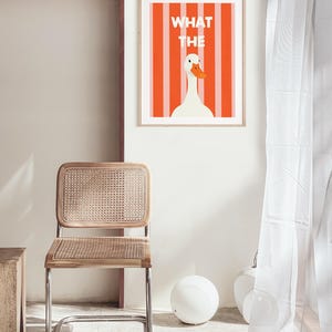 May include: A framed art print with a white goose and orange beak set against orange and white stripes. The words "WHAT THE" are above the goose. A wooden chair with a woven seat and back is in the foreground. The chair has a chrome frame.