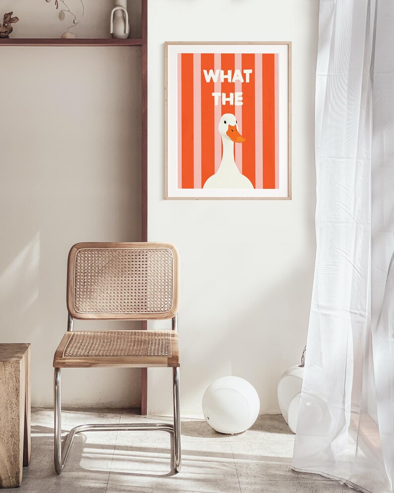 May include: A framed art print with a white goose and orange beak set against orange and white stripes. The words "WHAT THE" are above the goose. A wooden chair with a woven seat and back is in the foreground. The chair has a chrome frame.