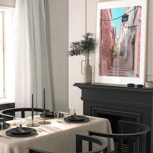 May include: A framed photograph of a narrow, cobblestone street in a European city. The street is lined with brick buildings and has a lamppost at the top of the stairs. The photo is hanging on a black fireplace mantel above a black fireplace.