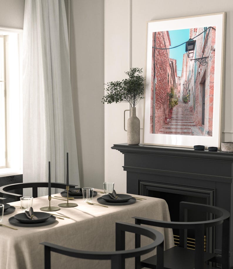 May include: A framed photograph of a narrow, cobblestone street in a European city. The street is lined with brick buildings and has a lamppost at the top of the stairs. The photo is hanging on a black fireplace mantel above a black fireplace.