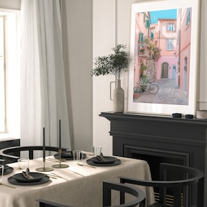 May include: A framed photograph of a narrow, cobblestone street in a European city. The buildings are painted in shades of pink and yellow, and there is a bicycle parked on the street. The photograph is hanging on a wall above a black fireplace.