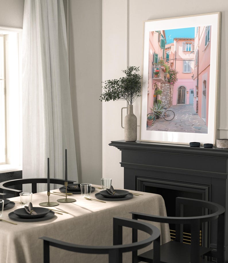 May include: A framed photograph of a narrow, cobblestone street in a European city. The buildings are painted in shades of pink and yellow, and there is a bicycle parked on the street. The photograph is hanging on a wall above a black fireplace.