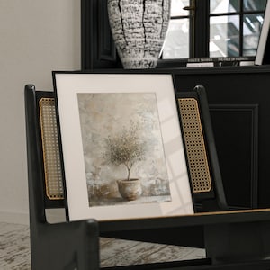 May include: A framed print of a small olive tree in a terracotta pot, set against a textured beige background. The print is displayed on a black chair with a woven cane back.