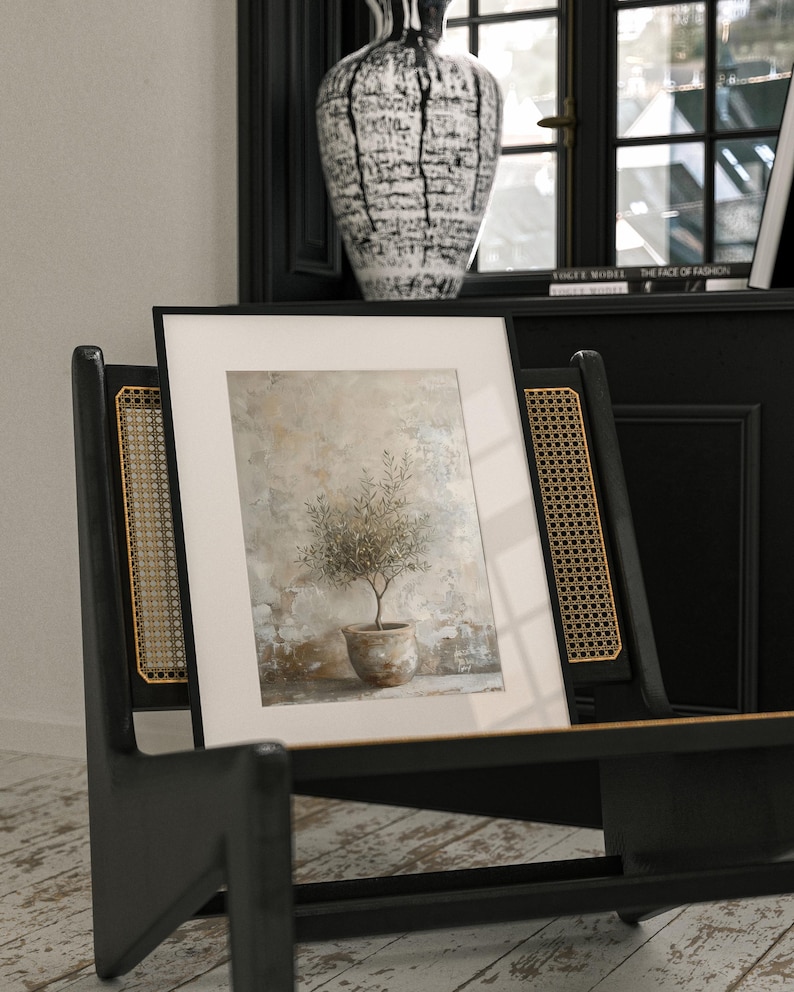 May include: A framed print of a small olive tree in a terracotta pot, set against a textured beige background. The print is displayed on a black chair with a woven cane back.