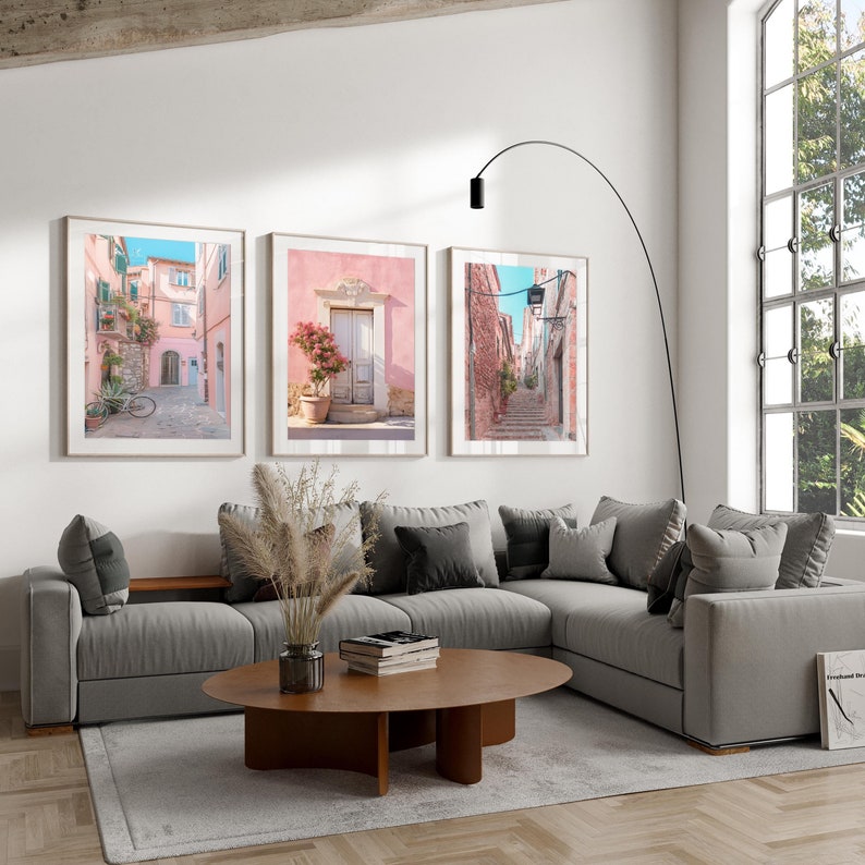 May include: Three framed photographs of Italian streets with pink buildings and cobblestone streets. The photos are hanging on a white wall in a living room with a gray sectional sofa and a wooden coffee table.