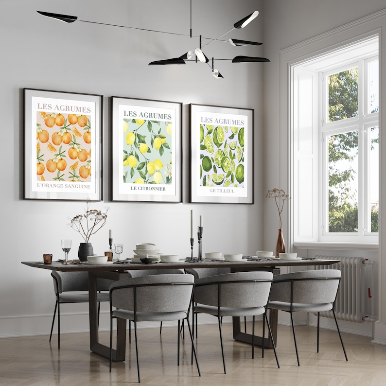 May include: Three framed prints with a white background and a colorful illustration of citrus fruits. The prints are titled "Les Agrumes" and each features a different type of citrus fruit: orange, lemon, and lime. The prints are hanging on a white wall above a wooden dining table with six chairs.