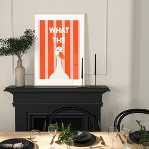 May include: Framed art print with a white goose and orange beak set against orange and white stripes. The text "WHAT THE" is above the goose. The print is displayed above a dining table setting with black plates and glasses.
