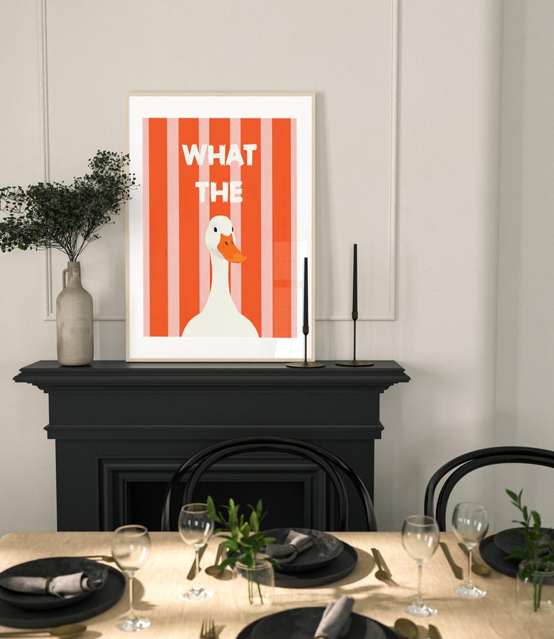 May include: Framed art print with a white goose and orange beak set against orange and white stripes. The text "WHAT THE" is above the goose. The print is displayed above a dining table setting with black plates and glasses.