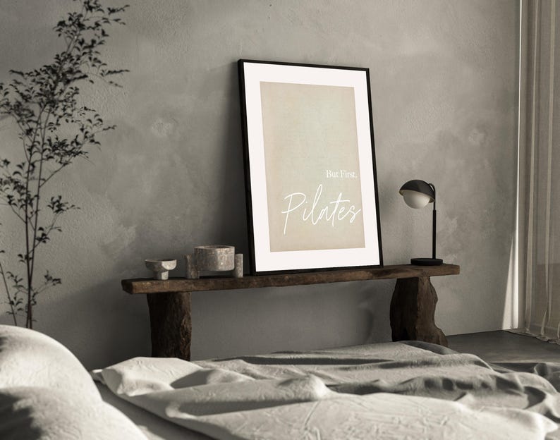 May include: A framed print with the words "But First, Pilates" in a soft, elegant script. The print is displayed on a rustic wooden bench, alongside decorative bowls and a small lamp. The scene is set in a neutral-toned room.