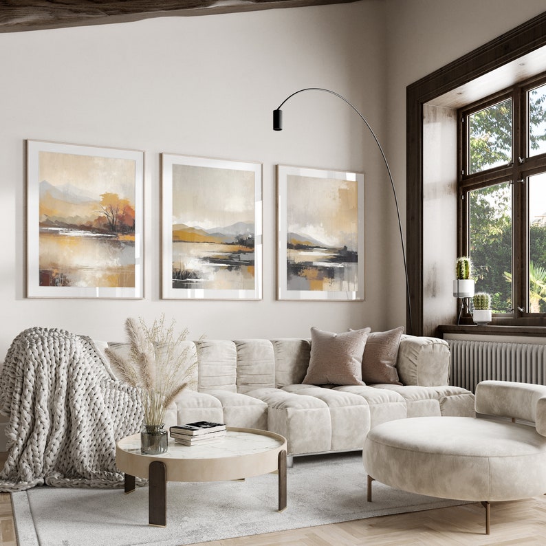 May include: A living room interior with a white sofa, a round coffee table, and three framed landscape paintings on the wall. The sofa has a light brown throw blanket and a light brown pillow. The coffee table has a white surface and a brown base. The paintings are all in the same style and depict a landscape with mountains, water, and trees. The room has a large window with a view of a green lawn and trees.