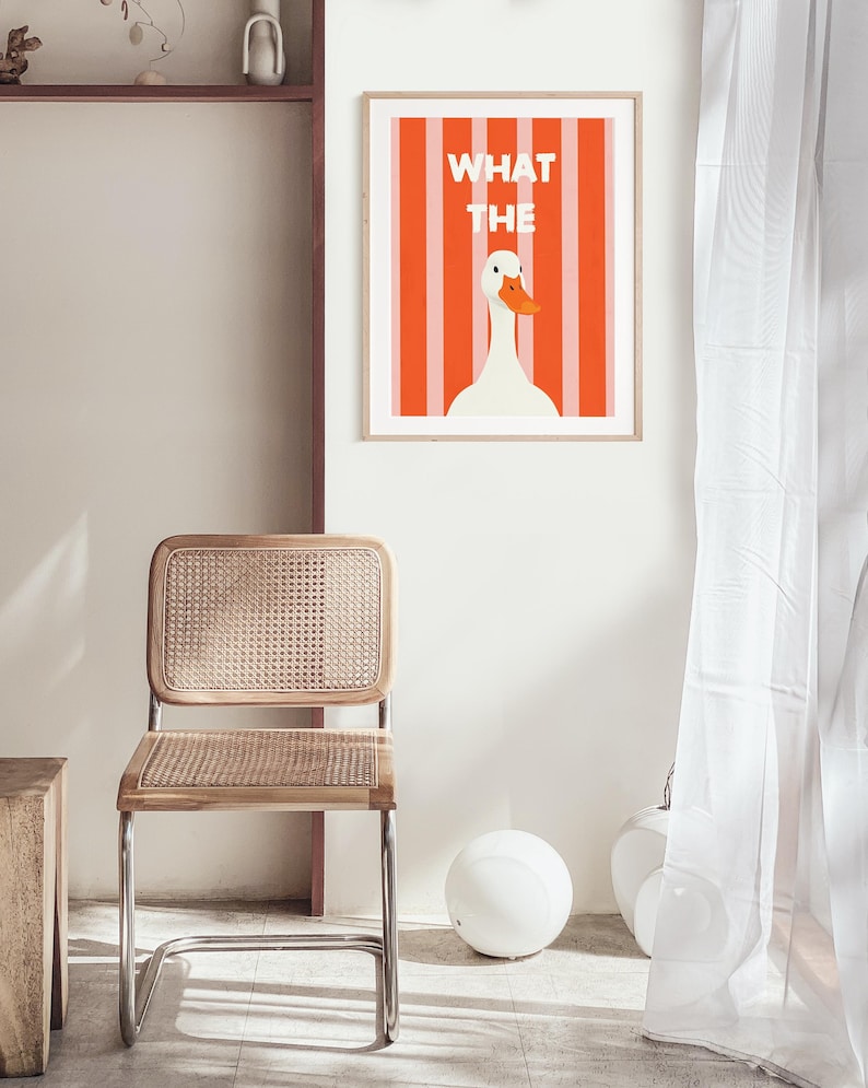 May include: A framed art print with a white goose and orange beak set against orange and white vertical stripes. The text "WHAT THE" is above the goose. A wooden chair with a woven seat and back is in the foreground. The chair has a chrome frame.