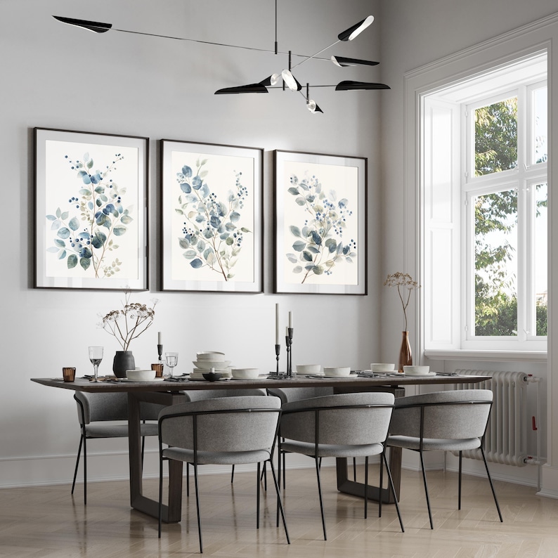 May include: A dining room with a wooden table and six chairs. The table is set with plates, glasses, and cutlery. Three framed watercolour paintings of blue and green leaves hang on the wall above the table.