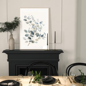 May include: A framed watercolour painting of a blue and green floral branch with a white background. The painting is hanging above a black fireplace mantel with a table set with black and gold place settings.