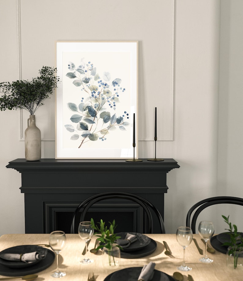 May include: A framed watercolour painting of a blue and green floral branch with a white background. The painting is hanging above a black fireplace mantel with a table set with black and gold place settings.