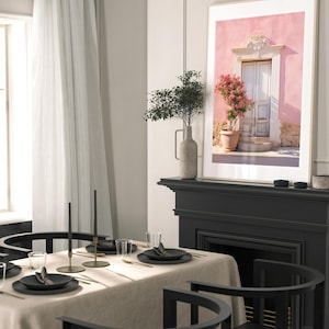 May include: A framed photograph of a pink building with a white door and a potted pink flower bush. The building has a white trim around the door and windows. The photo is hanging on a wall above a black fireplace.