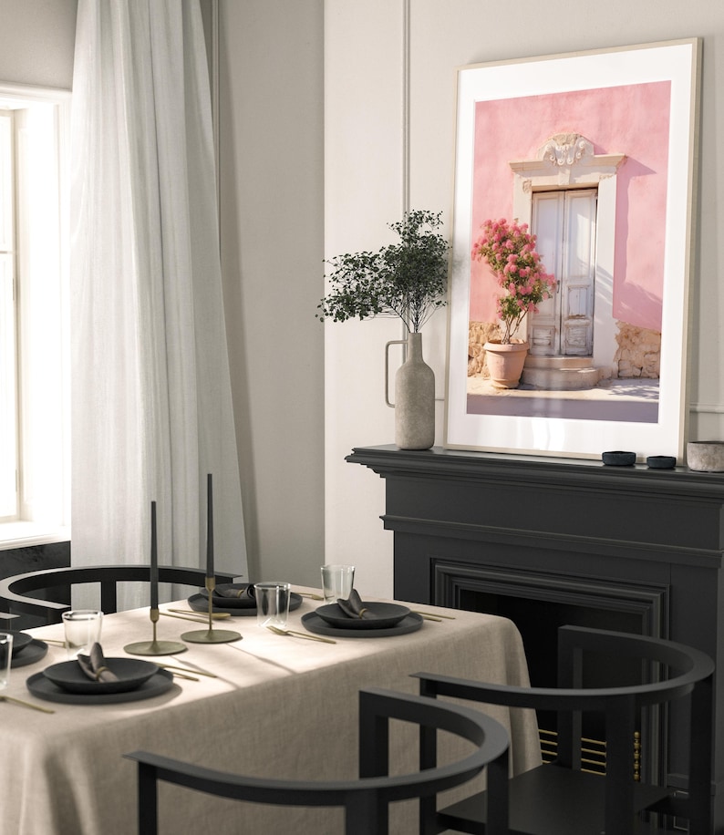 May include: A framed photograph of a pink building with a white door and a potted pink flower bush. The building has a white trim around the door and windows. The photo is hanging on a wall above a black fireplace.