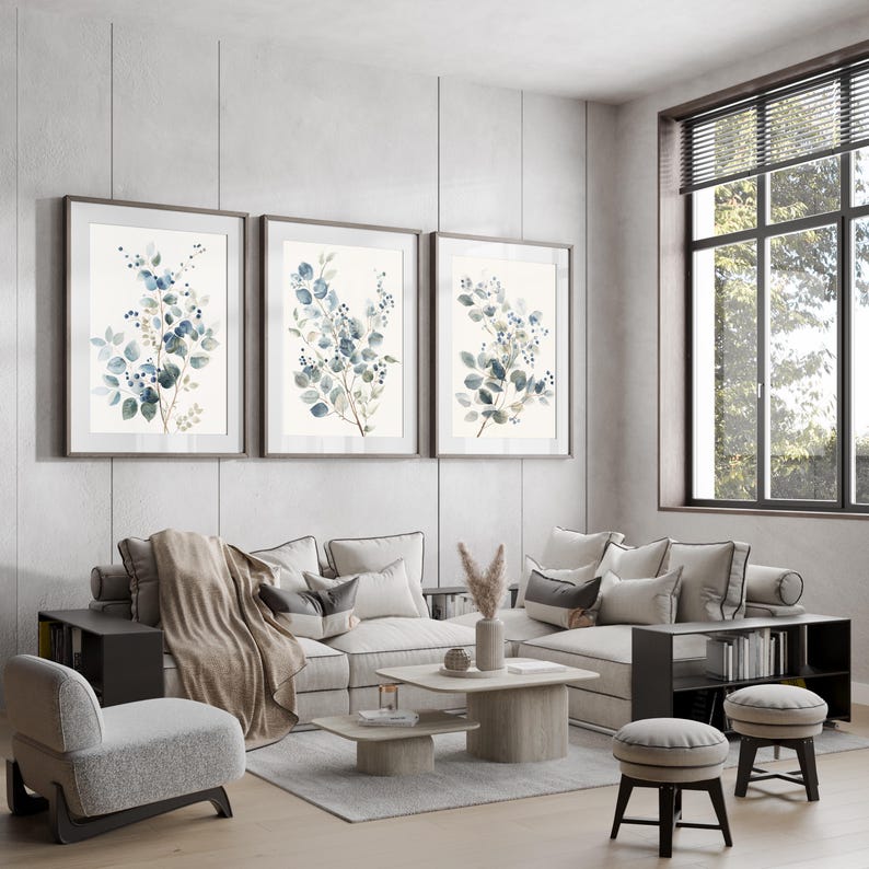 May include: A living room interior with a grey sectional sofa, three framed watercolour prints of blue and green foliage, a coffee table, and two grey and brown ottomans.