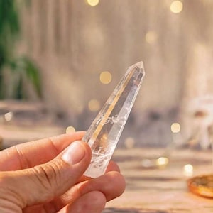 May include: A clear, pointed quartz crystal wand with a hexagonal shape and visible internal inclusions. The crystal is transparent and colourless, featuring a smooth, polished surface that tapers to a sharp point at the top end of the stone.