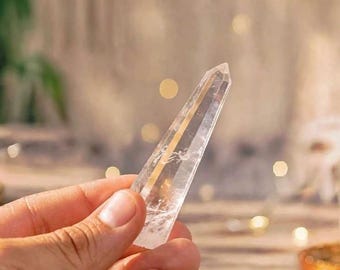 May include: A clear, pointed quartz crystal wand with a hexagonal shape and visible internal inclusions. The crystal is transparent and colorless, featuring a smooth, polished surface that tapers to a sharp point at the top end of the stone.